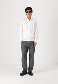 Weißes langärmliges Hemd mit Knöpfen, graue maßgeschneiderte Hose und schwarze klobige Schuhe. Person trägt schwarze Sonnenbrille. Minimalistisches Design, neutrale Farben.