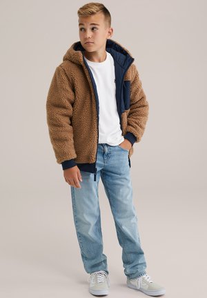 Veste en polaire beige avec une capuche, doublure navy et poignets côtelés ; associée à un t-shirt blanc et un jean délavé, complétée par des baskets grises.