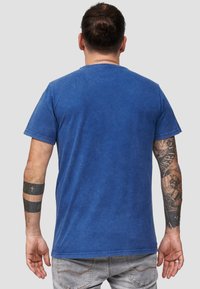 Sininen lyhythihainen t-paita, jossa on rento malli, pyöreä kaula-aukko ja pehmeä pinta. Näkyvät tatuoinnit käsissä ja farkut.