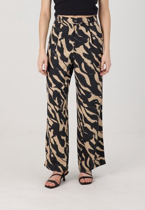 Kvinde iført beige vidde bukser med sort abstrakt print, sort cropped top og sorte rem-sandaler med hæl, stående mod en ensfarvet baggrund.