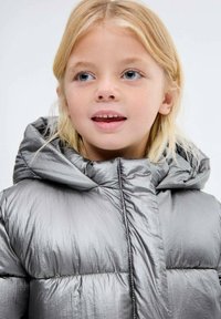 Malé dítě s blond vlasy, oblečené v lesklé stříbrné bundě s kapucí, dívající se mírně na stranu s lehce pootevřenými ústy.
