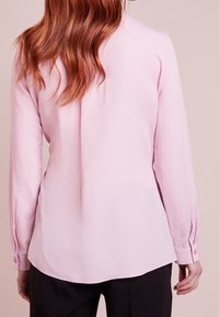 Vrouw met lang rood haar draagt een lichtroze blouse met lange mouwen en zwarte broek, gezien van de achterkant tegen een effen achtergrond.