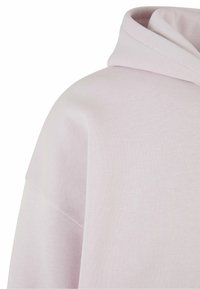 Felpa rosa chiaro con una texture morbida, dotata di un ampio cappuccio e vestibilità rilassata. Il tessuto appare liscio con minime cuciture visibili.