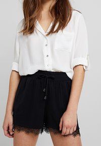 Vit kortärmad skjorta med knappar och ficka, kombinerad med svarta elastiska shorts med spetsdetaljer vid fållen.