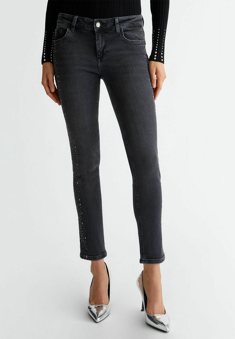 Femme portant un jean skinny noir avec de petits clous argentés le long des coutures latérales et des chaussures à talons hauts pointues argentées, debout.
