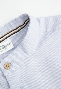 Jasnoniebieska koszula z kołnierzykiem o teksturowanym splocie, z drewnianym guzikiem i paskowanym wewnętrznym kołnierzem w kolorach brązowym, granatowym i białym.