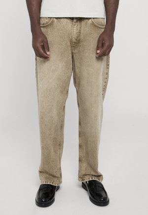 Person trägt beige Acid-Wash-Jeans, weißes Hemd und schwarze glänzende Loafers, steht vor einem schlichten hellgrauen Hintergrund.