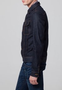 Mörk denimjacka med figurnära design, guldfärgade knappar, två framfickor och kontrasterande sömmar. Bärs med denimjeans.