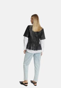 Schwarzes Kunstleder-Oberteil mit kurzen Ärmeln und volantiertem Saum, über einem weißen Hemd getragen; hellblaue weite Jeans und schwarze Slipper.