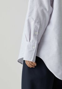 Camicia a righe viola chiaro e bianco con maniche lunghe. Presenta polsini con bottoni e orlo curvo. Indossata su pantaloni scuri.