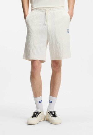 Mand iført teksturerede off-white snørebukser med sidelommer, hvide sokker med blåt logo og cremefarvede sneakers med sorte snørebånd.