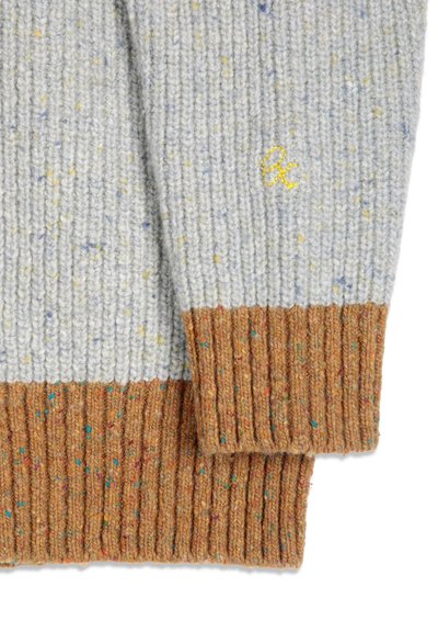 Gros plan sur la manche d'un pull gris en maille avec un bord-côtes marron et un petit logo jaune brodé.