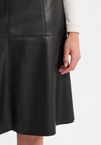 Zwarte leren rok met een uitlopende vorm, gladde textuur en nette stitching. Hand op de taille geplaatst, waardoor de stofdetails zichtbaar zijn.