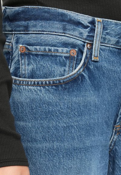 Close-up of blue denim jeans pocket with visible stitching, copper rivets, and a black ribbed sleeve resting near the waist.