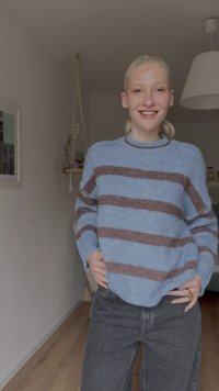 Pull tricoté bleu clair avec des rayures horizontales marron, col rond et poignets côtelés, associé à un jean gris foncé à jambes larges.