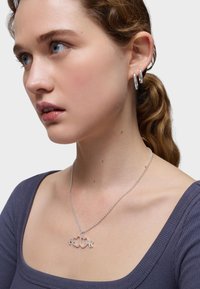 Collar de plata con corazones entrelazados y flechas, llevado sobre piel desnuda. La modelo lleva una blusa de textura púrpura y aretes de aro minimalistas.