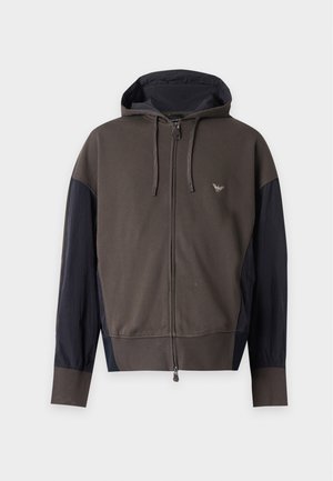 Jeune homme debout portant un sweat à capuche zippé marron foncé et noir, un t-shirt blanc avec un imprimé graphique, un pantalon noir ample et des baskets noires à semelles blanches.