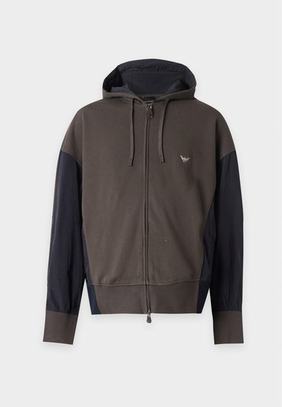 Sweat à capuche zippé marron foncé et noir, manches longues, capuche avec cordons et petit emblème sur le côté gauche de la poitrine.
