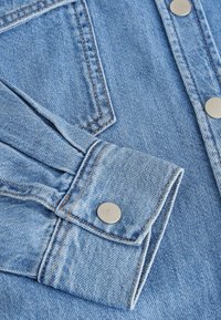 Camisa de denim azul clara con puños abotonados, que presenta un contraste de costuras naranjas y botones a presión de metal en la parte delantera.