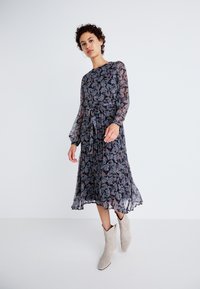 Φλοράλ μακρυμάνικη, ροή φόρεμα με σκοτεινό λουλούδι σε διάφανη ύλη, δεμένο στη μέση και πλισέ φούστα, σε συνδυασμό με ανοιχτό γκρι μποτάκια μέχρι τον αστράγαλο.