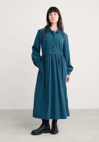 Abito in velluto a coste color teal con colletto abbottonato, maniche lunghe e vita arricciata. Presenta una tasca sul petto e arriva fino alle caviglie. Stivali neri alla caviglia.
