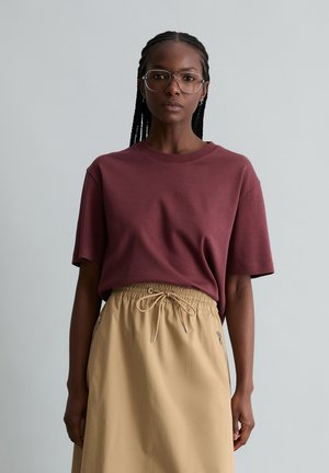 Kobieta z warkoczem i okularami, ubrana w bordową koszulkę z krótkim rękawem wsuniętą w beżową spódnicę ze sznurkiem, na jednolitym tle.