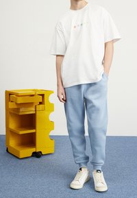 Bílý bavlněný tričko s vícebarevnou grafikou; světle modré tepláky; bílé tenisky. Žlutá úložná skříň v pozadí, texturovaný povrch.