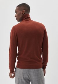 Rūsgans krāsas turtlenecks džemperis, izgatavots no mīkstas auduma, ar garām piedurknēm un ribotu apkakli, kombinēts ar pelēkām trijstūrainām biksēm.