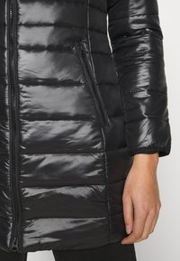 Chaqueta negra acolchada con un acabado brillante, que presenta secciones acolchadas horizontales y un bolsillo lateral con cremallera. Los puños son elásticos.