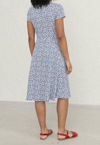 Modré a bílé květinové šaty s krátkými rukávy, přiléhavým živůtkem a rozšířenou sukní, vyrobené z lehkého materiálu. Červené sandály doplňují vzhled.