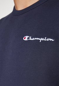 T-shirt bleu marine avec logo Champion brodé en blanc sur la partie supérieure de la poitrine, avec un emblème rouge et blanc.