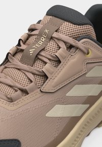 Ανοιχτό καφέ αθλητικό παπούτσι Adidas Terrex με μπεζ ρίγες, επάνω μέρος από δίχτυ, επενδεδυμένο γιακά και λεπτομερείς θηλιές για τα κορδόνια σε λευκό φόντο.