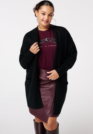 Femme aux cheveux tressés portant de grandes boucles d'oreilles en forme de créole, un cardigan noir, un t-shirt bordeaux "LOVER", une jupe en cuir et des bottes assorties, souriant doucement.