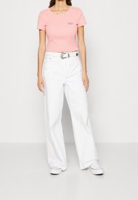 T-shirt di cotone rosa chiaro con scollatura rotonda e piccolo logo sul petto, abbinato a jeans bianchi a gamba larga e cintura argentata.