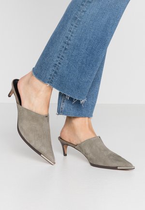 Füße mit grauen, spitz zulaufenden Wildleder-Mules mit kleinen Absätzen, kombiniert mit blauen Jeans mit ausgefranstem Saum auf weißem Boden.