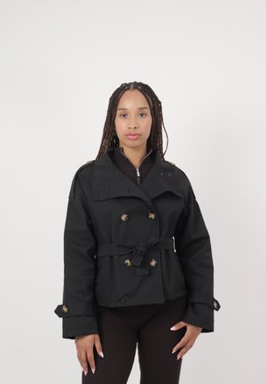 Kvinde med lange fletninger står vendt fremad iført sort trenchcoat med bælte og guldfarvede knapper over mørke bukser mod en neutral baggrund.