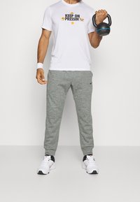 Vit t-shirt med kort ärm och svart grafik "KEEP ON PRESSIN'", tillsammans med gråa smalare joggers och vita sneakers med svarta detaljer.