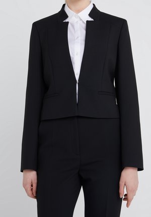 Blazer noir cropped avec un design structuré, doté d'un col haut, de poches avant et d'une ouverture à l'avant. Porté sur une chemise blanche.
