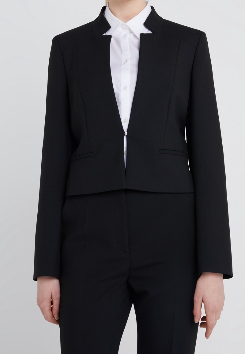 Blazer corto nero con un design strutturato, caratterizzato da un colletto alto, tasche frontali e un'apertura frontale. Indossato sopra una camicia bianca.