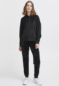 Tuta in velour nero con pullover a mezza zip e tasca a marsupio, pantaloni affusolati. Abbinato a sneakers sportive bianche.