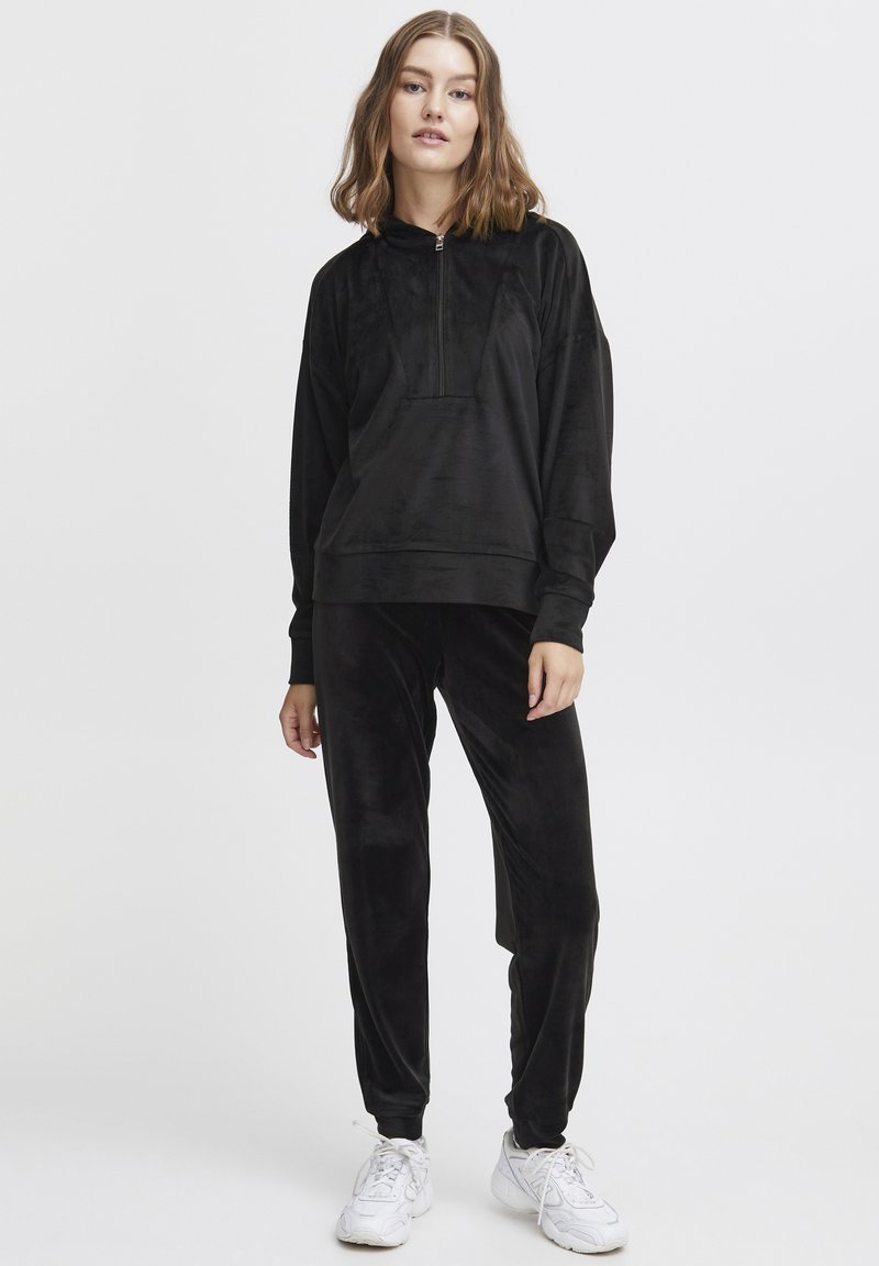 Tuta in velour nero con pullover a mezza zip e tasca a marsupio, pantaloni affusolati. Abbinato a sneakers sportive bianche.