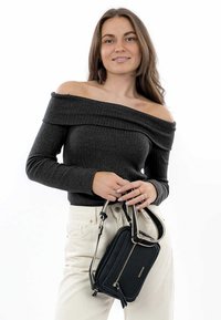 Černá texturovaná crossbody taška s dvojitým zipem a krátkým popruhem, držena osobou v šedém žebrovaném topu s odhalenými rameny a krémových kalhotách.