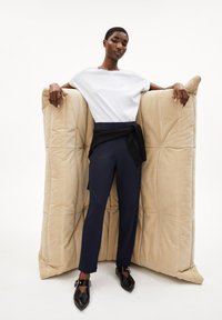 T-shirt bianco, pantaloni fitted blu scuro e scarpe nere a punta; in piedi tra due grandi cuscini beige di peluche con dettagli imbottiti.