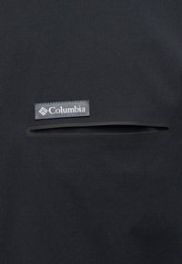 Μαύρο ύφασμα με οριζόντια τσέπη με φερμουάρ και γκρι ετικέτα με την ένδειξη "Columbia" ραμμένη πάνω από το άνοιγμα της τσέπης.