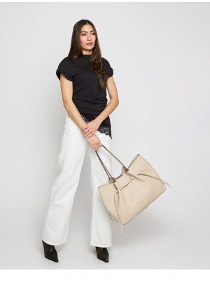 Woman wearing black top and white pants holding large beige handbag, standing against plain gray background.