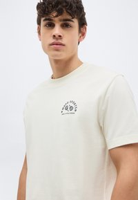 Νεαρός άνδρας με σγουρά μαλλιά που φοράει ένα κρεμ t-shirt με ένα μικρό λογότυπο "Mood Sports Marc O’Polo Denim" στο στήθος.