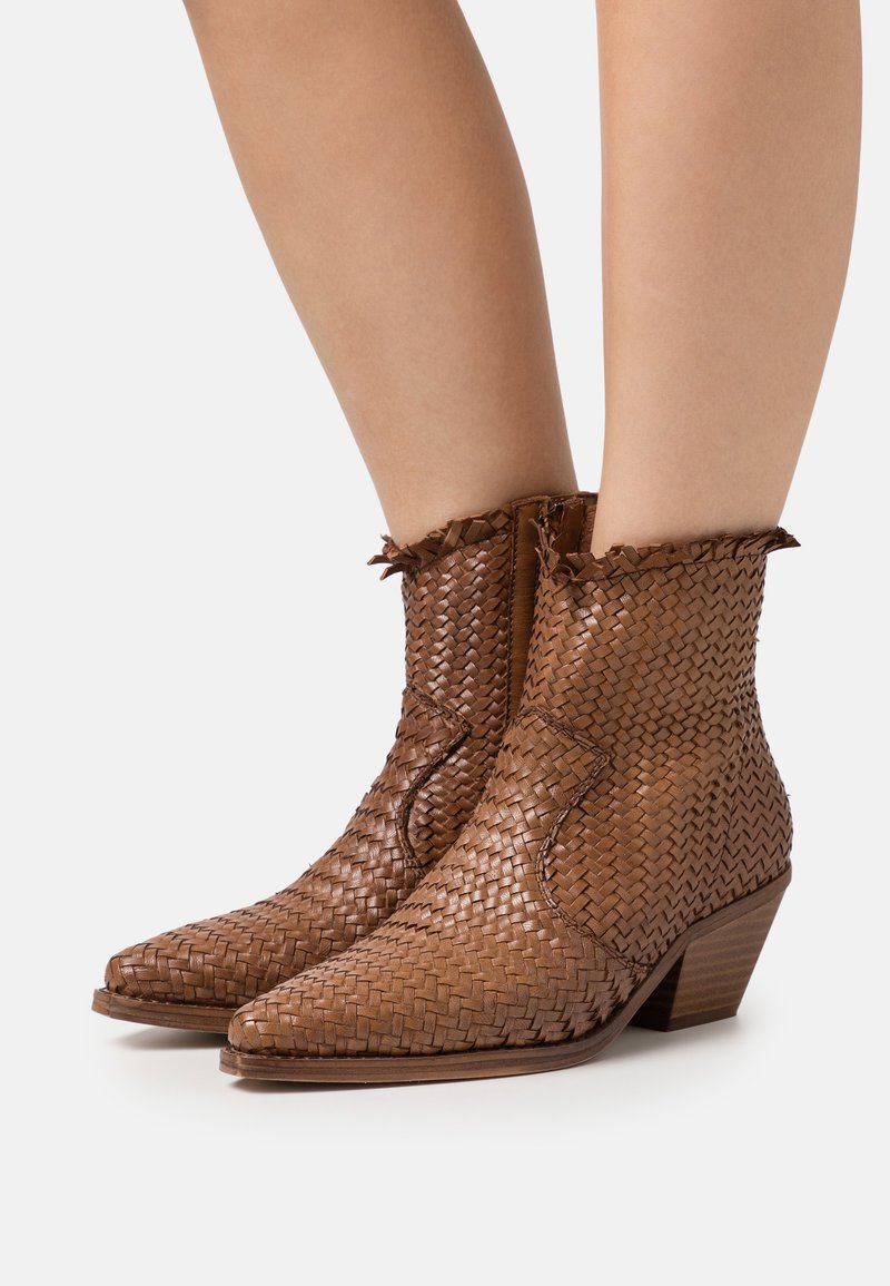 Botas de tobillo de cuero trenzado marrón con punta afilada, borde superior deshilachado y un tacón bajo de madera. Superficie texturizada con patrones distintivos.