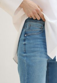 Svetlo modri jeans z oprijetim krojem, vidnim šivi in sprednjimi žepi. Bela majica delno prekrije pas.