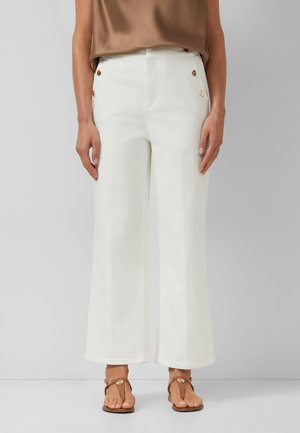 Białe spodnie o szerokich nogawkach z złotymi guzikami na biodrach, noszone z beżowym topem bez rękawów i brązowymi sandałami z ozdobami z muszli.