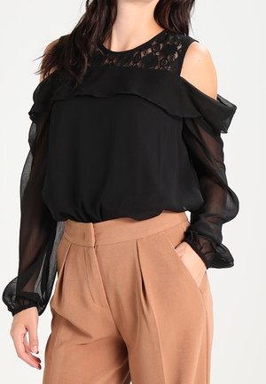 Blouse noire dénudée des épaules en tissu léger, ornée de dentelle et de détails volantés au niveau du col et des manches.